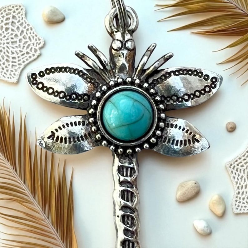 May include: Silver-toned dragonfly pendant with a turquoise-colored stone center. The dragonfly's wings have intricate detailing, and the pendant hangs from a silver chain. The pendant is a jewelry item.