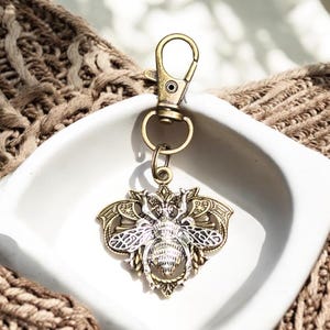 May include: A gold and silver bee keychain with intricate details. The bee is surrounded by a decorative frame with wings. The keychain has a lobster clasp.