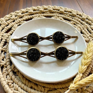 May include: Four hair barrettes with a circular black stone design set in a gold-toned metal frame. The barrettes are arranged on a white plate, which is placed on a woven placemat. The barrettes are a stylish hair accessory.