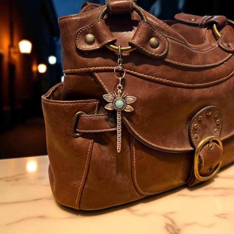 May include: A brown leather handbag with a decorative dragonfly charm. The handbag features brass hardware, including a buckle and ring details. The dragonfly charm is silver-toned with a turquoise stone center.