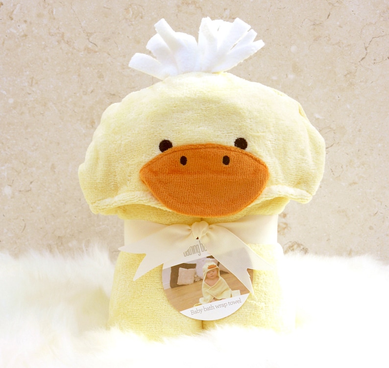 Peut inclure: Une serviette &agrave; capuche jaune avec un visage de canard et une cr&ecirc;te blanche et moelleuse. La serviette est envelopp&eacute;e et nou&eacute;e avec un ruban blanc. L'&eacute;tiquette indique "Serviette de bain enveloppante pour b&eacute;b&eacute;".