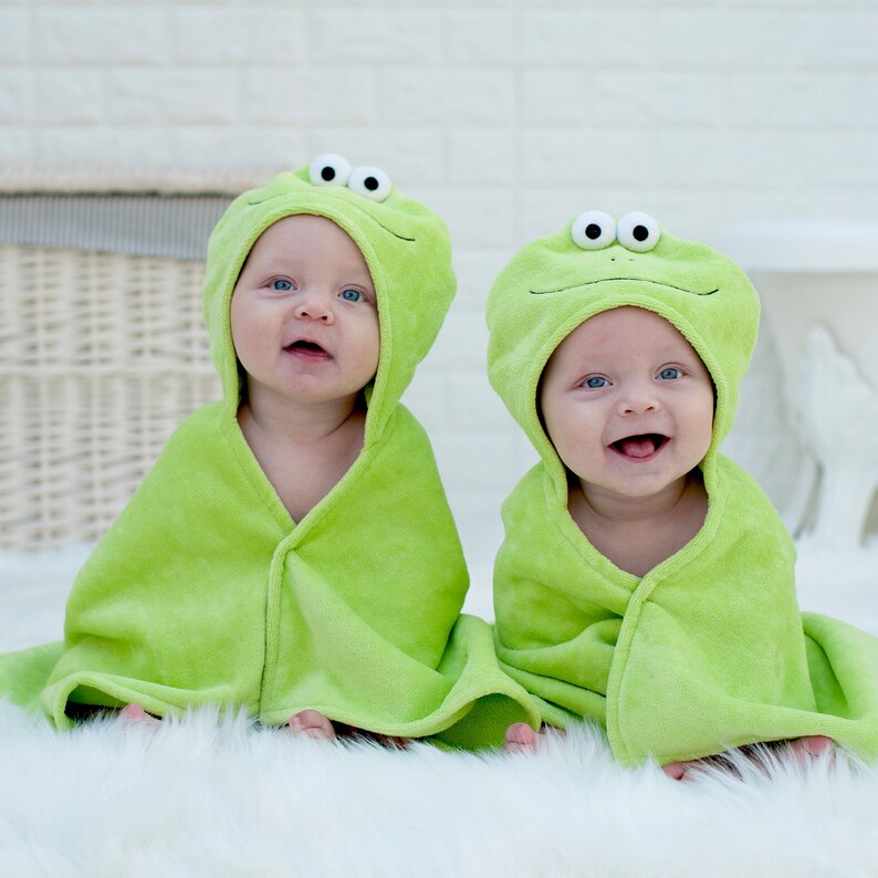Puede incluir: Dos beb&eacute;s con toallas con capucha verde con caras de rana. Las toallas tienen ojos grandes y redondos y una amplia sonrisa.