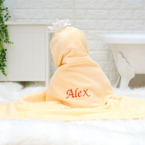 Peut inclure: Une serviette &agrave; capuche jaune p&acirc;le avec une fleur blanche sur la capuche. La serviette est brod&eacute;e du nom "Alex" en fil rouge.