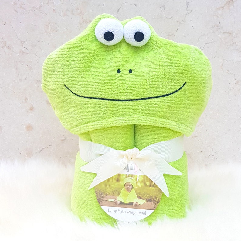 Puede incluir: Toalla con capucha verde con cara de rana, ojos grandes y una sonrisa. La toalla est&aacute; envuelta y atada con una cinta blanca. La etiqueta dice "Toalla envolvente para ba&ntilde;o de beb&eacute;".