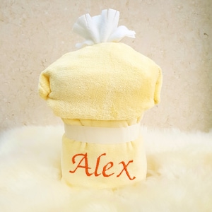Peut inclure: Serviette &agrave; capuche jaune avec une capuche blanche et un ruban blanc. La serviette a le nom "Alex" brod&eacute; sur le devant en fil orange.