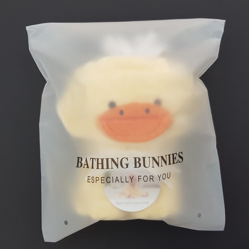 Peut inclure: Une serviette &agrave; capuche en forme de canard jaune avec un bec orange et des yeux noirs. La serviette est emball&eacute;e dans un sac en plastique transparent avec le texte "BATHING BUNNIES ESPECIALLY FOR YOU" et "Baby bath wrap towel".