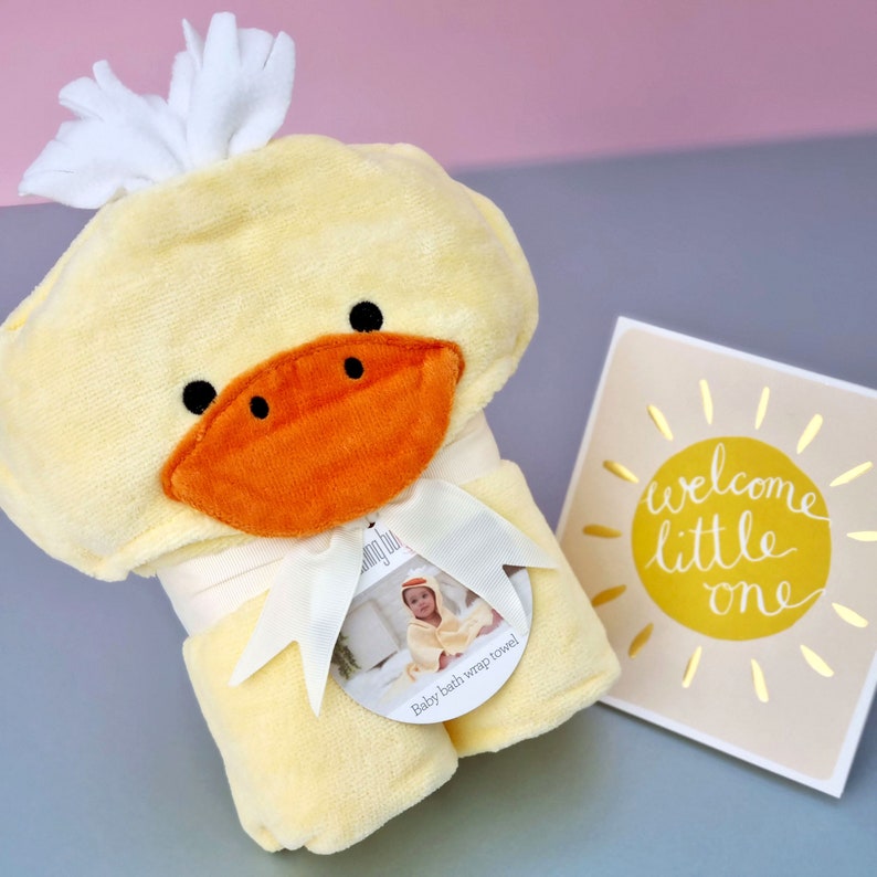 Peut inclure: Serviette &agrave; capuche en forme de canard jaune avec un n&oelig;ud blanc et une &eacute;tiquette indiquant "Baby bath wrap towel!" La serviette est roul&eacute;e et plac&eacute;e &agrave; c&ocirc;t&eacute; d'une carte qui dit "Welcome little one" avec un graphique de soleil jaune.