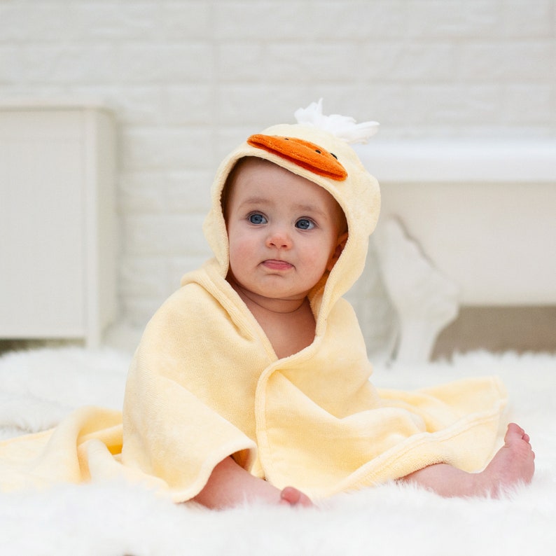 Peut inclure: Une serviette &agrave; capuche jaune avec un visage de canard sur la capuche. Un b&eacute;b&eacute; porte la serviette et est assis sur une surface blanche et moelleuse.