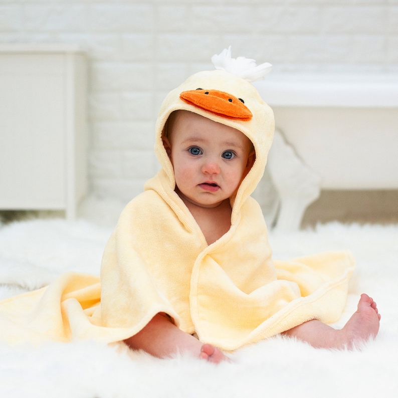 Peut inclure: Une serviette &agrave; capuche jaune avec un visage de canard sur la capuche. Un b&eacute;b&eacute; porte la serviette et est assis sur une surface blanche et moelleuse.