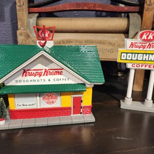 May include: A miniature Krispy Kreme shop and sign. The shop has a green roof, white trim, and red and yellow accents. The sign reads "Doughnuts Coffee". A small Christmas tree is in front of the shop.