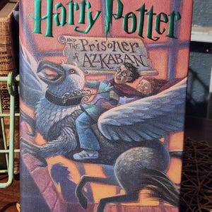 May include: A hardcover book titled "Harry Potter and the Prisoner of Azkaban" by J.K. Rowling. The cover features a colorful illustration of Harry Potter riding a Hippogriff, with the title in green lettering.