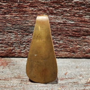 May include: A vintage brass shoehorn with a curved handle and a wide, rounded end. The shoehorn has a tarnished, aged appearance, with visible scratches and wear. The background is a weathered wooden surface.