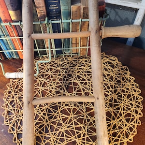 May include: A vintage wooden clothes drying rack with a handle, featuring a ladder-like design. The rack is light brown and sits on a woven placemat. A wire basket filled with old books is in the background.