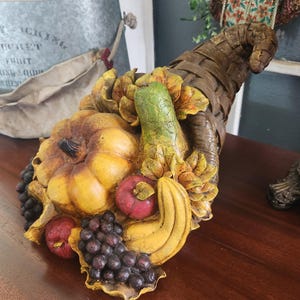 May include: A decorative cornucopia overflowing with a variety of faux fruits and vegetables. The arrangement includes a yellow pumpkin, green pear, red apples, purple grapes, and yellow bananas. The horn is woven brown, and the base is a golden yellow.