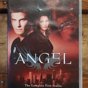May include: DVD case for the first season of the TV show "Angel". The case features a red background with a cityscape and the show's title in white. The text "The Complete First Season" and "6-Disc Set" are also visible.