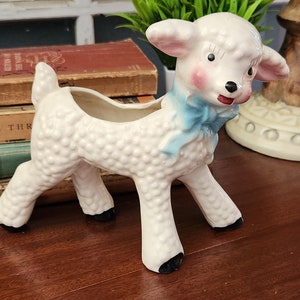 May include: White ceramic lamb planter with a blue bow. The lamb has a textured surface and black eyes. The planter has a hole in the top for drainage.