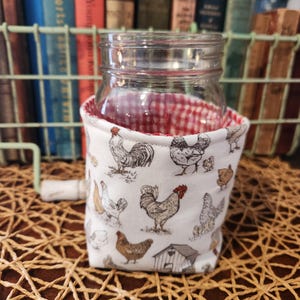 May include: A clear glass jar with a red and white checkered lining is encased in a white fabric cozy. The cozy features a repeating pattern of chickens, roosters, and a small chicken coop. The jar sits on a woven mat.