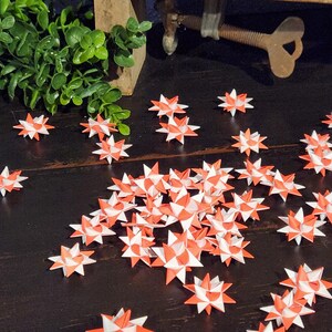 May include: A collection of origami stars in red and white scattered on a dark wooden surface. The star-shaped paper ornaments are folded with intricate detail. Green foliage and a metal tool are visible in the background.
