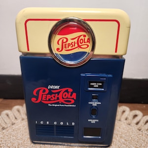 May include: A vintage-style Pepsi-Cola vending machine replica featuring a cream-colored top section with a circular Pepsi logo, a dark blue body with the red Pepsi-Cola logo, and text reading Drink, The Original Pure Food Drinks, and Ice Cold.