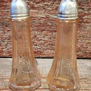 May include: Two vintage, peach-colored glass salt shakers with silver-colored metal tops. The glass shakers have a floral pattern and a hexagonal base. The shakers are displayed on a wooden surface.