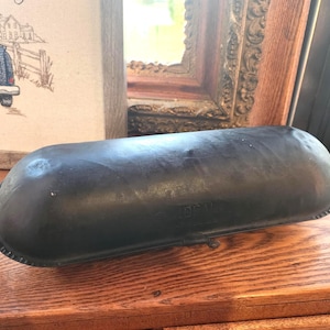 May include: A dark, oval-shaped, antique wooden dough bowl. The bowl has a textured surface and a decorative rim. It sits on a wooden surface, suggesting a rustic or farmhouse aesthetic.