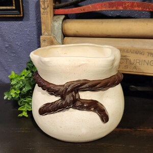 May include: Cream-colored ceramic vase with a dark brown rope design around the middle. The vase has a rounded body and a slightly flared top. The rope is intricately detailed, adding a rustic touch to the piece.