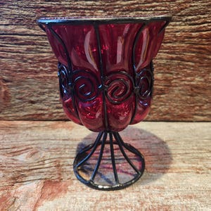May include: A ruby red glass votive candle holder with a black metal filigree design. The holder has a rounded bowl shape and sits on a black metal base. The metalwork features decorative swirls.