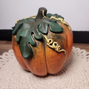 May include: A decorative pumpkin figurine featuring a textured orange body with deep vertical ridges. The top is adorned with a dark green sculpted leaf and a small, curled yellow vine detail. A dark brown stem sits at the very center of the pumpkin.