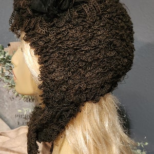 May include: A brown crocheted hat with a black fabric flower accent. The hat is on a mannequin head with blonde hair.