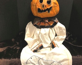 May include: A handmade Halloween doll with a white dress and a pumpkin head. The pumpkin has a carved face and a brown stem. The doll is sitting on a brown surface and has a black bird embroidered on its dress.