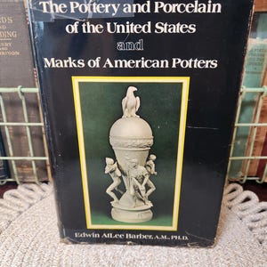 May include: A vintage book titled "The Pottery and Porcelain of the United States and Marks of American Potters." The cover is black with white text and a central image of a white sculpture of a vase with figures and an eagle.