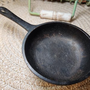 May include: A small, black cast iron skillet with a long handle and a hole for hanging. The pan has a round cooking surface and is sitting on a woven mat. The skillet is likely used for cooking small portions.