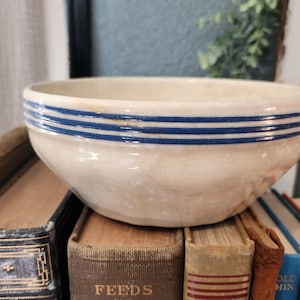 May include: Cream-colored ceramic bowl with three horizontal blue stripes around the rim. The bowl is sitting on top of a stack of vintage books with various colors and textures. A classic kitchenware item.