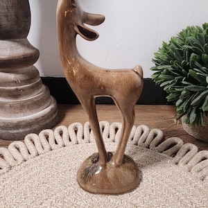 May include: A standing ceramic deer figurine with a glossy brown glaze. The deer has an elongated neck, a small tail, and is positioned on a circular base that features a small molded pinecone detail at the feet of the animal figure.