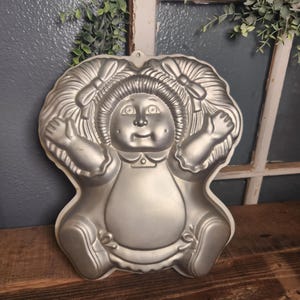 May include: A silver-colored metal cake pan shaped like a doll with yarn-style hair in pigtails, a round face, outstretched arms, and legs tucked underneath. The mold features embossed details including facial features, hair bows, and clothing.