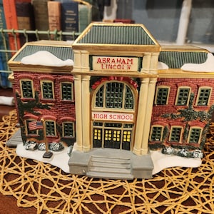 May include: A decorative miniature schoolhouse with the text "ABRAHAM LINCOLN HIGH SCHOOL". The building is made of red brick with green trim and a gold accent. An American flag stands in front.