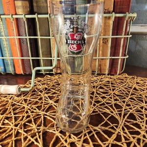 May include: A clear glass beer boot with a gold rim and the Beck's logo. The boot-shaped glass sits on a woven mat, with a wire basket of books in the background.