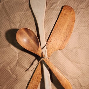 May include: A collection of wooden kitchen utensils, including a spoon, spatula, and turner, tied together with twine. The spoon is a rich brown color, while the spatula is a light gray. The turner and spoon are a lighter brown color.