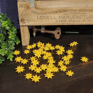 May include: A collection of small, yellow star-shaped objects scattered on a dark wooden surface. A vintage wooden sign with text is in the background, along with a green plant. The stars are the focal point.