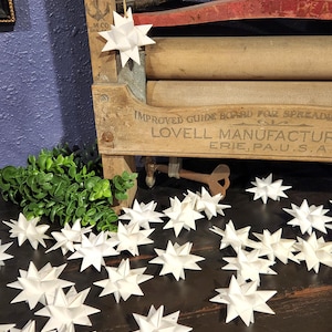 May include: White paper star ornaments scattered on a dark wooden surface. One star hangs from a gold string on a vintage wooden laundry machine. The machine has the text "IMPROVED GUIDE BOARD FOR SPREADING" and "LOVELL MANUFACTURING ERIE, PA. U.S.A."