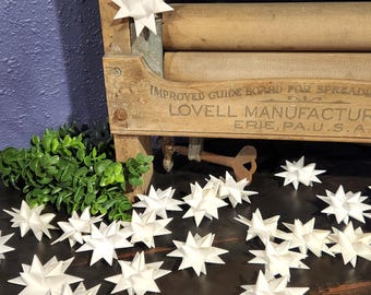 May include: White paper star ornaments scattered on a dark wooden surface. One star hangs from a gold string on a vintage wooden laundry machine. The machine has the text "IMPROVED GUIDE BOARD FOR SPREADING" and "LOVELL MANUFACTURING ERIE, PA. U.S.A."