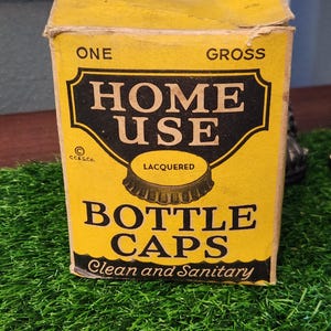 May include: A yellow cardboard box with black text that reads "One Gross Home Use Lacquered Bottle Caps Clean and Sanitary". The box has a picture of a bottle cap on it.
