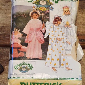 May include: A vintage Butterick sewing pattern for Cabbage Patch Kids outfits. The package features illustrations of children in angel and fairy costumes. The pattern includes iron-on transfers. The package is yellow and white.