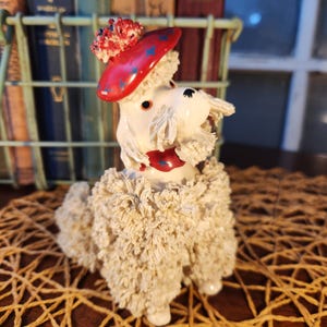 May include: A ceramic poodle figurine with a red beret decorated with blue stars and a matching red collar. The poodle has a white, textured coat and is posed on a woven mat. The figurine is a decorative item.