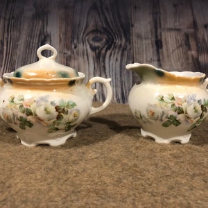 May include: A vintage white porcelain sugar bowl and creamer set with a floral design featuring white roses and green leaves. The set has a gold trim and a scalloped edge.