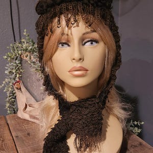 May include: A brown crocheted hat with a fringe and a black flower accent. The hat is worn on a mannequin with a brown crocheted scarf.