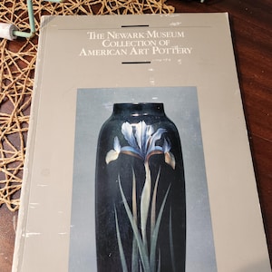 May include: A book titled "The Newark Museum Collection of American Art Pottery" with a dark blue vase illustration featuring a white and blue iris flower. The book is on a wooden surface.