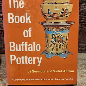 May include: An orange book cover with the title "The Book of Buffalo Pottery" in white text. The cover features a detailed illustration of a Buffalo Pottery vase. The book is by Seymour and Violet Altman.