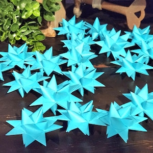 May include: A collection of bright turquoise paper star ornaments. Each star has multiple points and is folded from paper. The stars are scattered on a dark wooden surface, with greenery and a decorative object in the background.