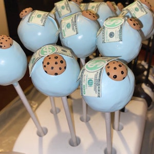May include: Light blue cake pops decorated with a chocolate chip cookie and a paper $100 bill. The cake pops are arranged in a white holder, with several visible in the image. The cookies are brown with dark brown chocolate chips.